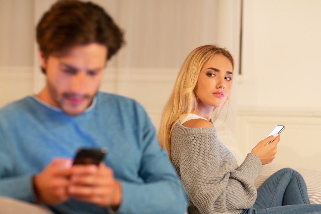 Wife Watching Husband Texting On Phone Suspecting Infidelity Sitting Indoor